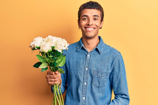 Fiatal afro-amerikai férfi kezében virágok látszó pozitív és boldog álló és mosolygó magabiztos mosollyal mutatja fogak  - Fotó, kép