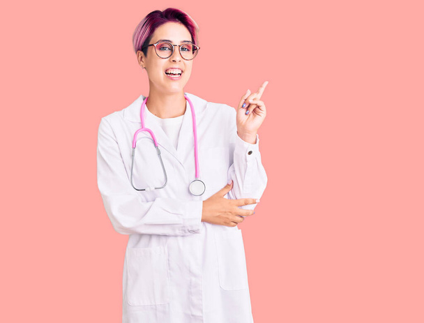 Młoda piękna kobieta w różowych włosach w mundurze lekarskim z dużym uśmiechem na twarzy, wskazując ręką i palcem na bok patrząc w kamerę.  - Zdjęcie, obraz