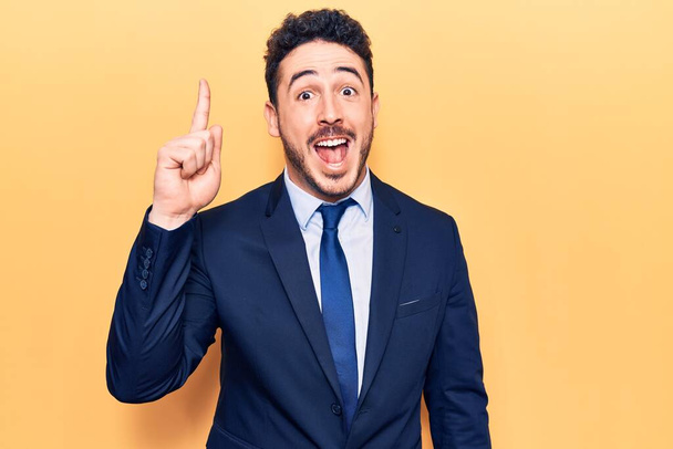 Junger hispanischer Mann im Anzug zeigt mit erhobenem Zeigefinger auf erfolgreiche Idee. aufgeregt und glücklich. Nummer eins.  - Foto, Bild