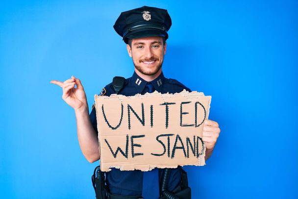 Молодой кавказский человек в полицейской форме, держащий вместе мы стоим знамя улыбаясь счастливо указывая рукой и пальцем в сторону  - Фото, изображение