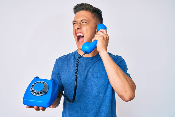 Junger hispanischer Junge, der ein altes Telefon in der Hand hält, wütend und wütend schreit und vor Wut schreit. Wut und aggressives Konzept.  - Foto, Bild