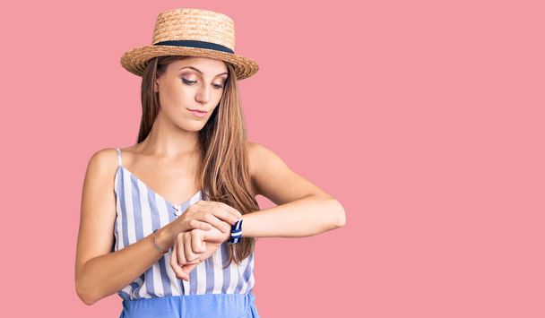 Νεαρή όμορφη ξανθιά γυναίκα φοράει καλοκαιρινό καπέλο ελέγχοντας την ώρα στο ρολόι του καρπού, χαλαρή και με αυτοπεποίθηση  - Φωτογραφία, εικόνα