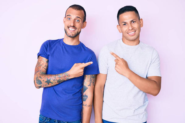 Nuori homo pari kaksi miestä yllään rento vaatteita hymyillen ja katsoen kameraa osoittaen kahdella kädellä ja sormella sivulle.  - Valokuva, kuva