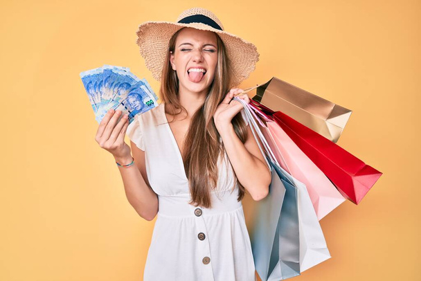 Joven chica rubia sosteniendo bolsas de compras y rands sudafricanos billetes que salen con la lengua feliz con expresión divertida.  - Foto, Imagen