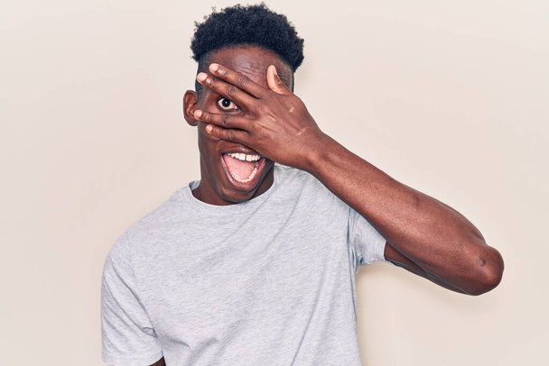 Mladý afroameričan v neformálním oblečení vykukuje v šoku, zakrývá obličej a oči rukou, dívá se prsty s rozpačitým výrazem.  - Fotografie, Obrázek