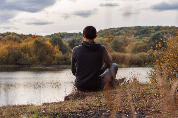 Un jeune homme est assis sur la rive de la rivière, dans la soirée nuageuse d'automne. Vue arrière - Photo, image