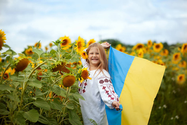 Ukrainisches lächelndes Mädchen in einem traditionellen bestickten Hemd mit einer gelb-blauen Flagge in den Händen auf einem Feld mit Sonnenblumen - Foto, Bild