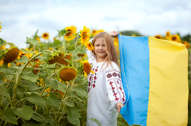 Ουκρανικό χαμογελαστό κορίτσι σε ένα παραδοσιακό κεντημένο πουκάμισο με κίτρινη-μπλε σημαία στα χέρια της σε ένα πεδίο με ηλιοτρόπια - Φωτογραφία, εικόνα