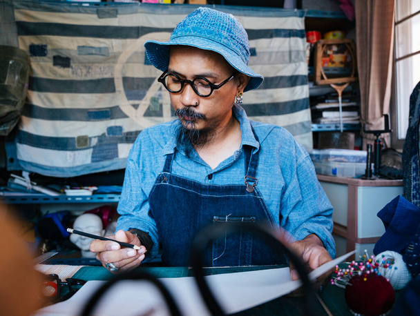 Ένας άντρας με μπλε ρούχα, καπέλο και ποδιά σκοπεύει να φτιάξει ο ίδιος τα παραδοσιακά ρούχα.. - Φωτογραφία, εικόνα