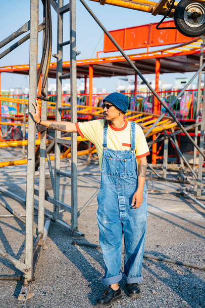 A man in yellow t-shirt and denim overalls standing in the abandoned amusement park in sunny day. - Photo, Image