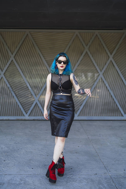 A cool young lady with blue hair, tattoos, and sunglasses, wearing a crop top, leather pencil skirt and red shoes posing in front of a garage - Foto, Imagen