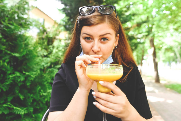 A maszkos lány koktélt iszik a kávézó nyári teraszán. Karantén. Új valóság. - Fotó, kép