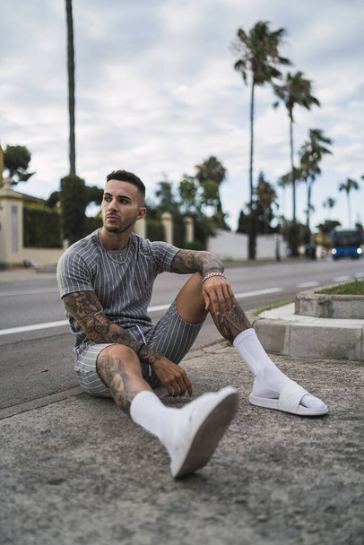 A vertical shot of an athletic young man with tattoos posing sitting on the street - Zdjęcie, obraz