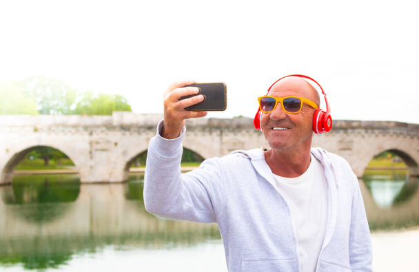 senior man takes a selfie and smiles listening to music with headphones - old man takes a selfie outdoor - concept about happy retirement - Photo, Image