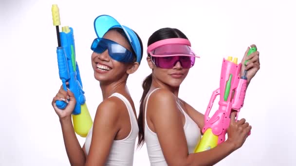 Portret dwóch pięknych Azjatek w różowej i niebieskiej czapce z daszkiem i okularach przeciwsłonecznych pozujących z pistoletami wodnymi na białym tle - film w zwolnionym tempie - Materiał filmowy, wideo