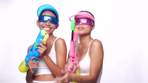 Retrato de dos hermosas mujeres asiáticas en gorra de visera de sol rosa y azul y gafas de sol posando con pistolas de agua sobre fondo blanco - Metraje, vídeo