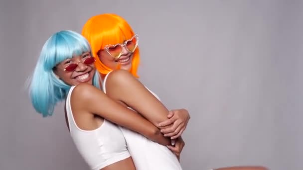Retrato de dos hermosas amigas asiáticas felices en pelucas y gafas de sol azules y naranjas brillantes posando sobre fondo gris - Imágenes, Vídeo