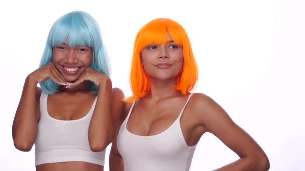 Portré két gyönyörű boldog ázsiai nők barátok fényes kék és narancssárga paróka pózol fehér háttér - Felvétel, videó