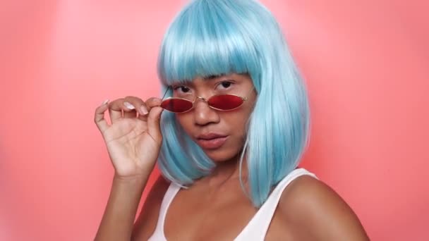 Portré gyönyörű boldog ázsiai nő fényes kék paróka és napszemüveg pózol rózsaszín háttér - Felvétel, videó