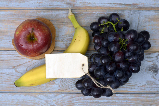 A bunch of fresh organic black grapes, banana, red apple, and a blank label tag on a wooden table - Fotoğraf, Görsel