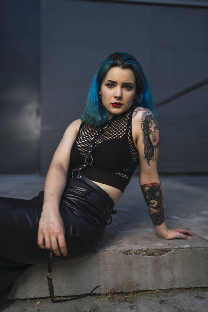 A cool young lady with blue hair, tattoos, wearing a crop top, leather pencil skirt, posing sitting on the ground - Foto, Imagen