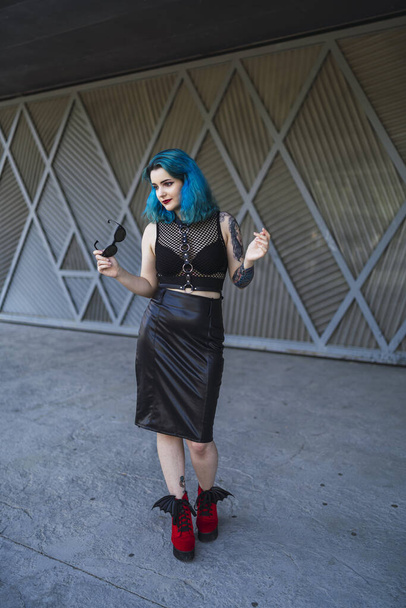 A cool young lady with blue hair, tattoos, and sunglasses, wearing a crop top, leather pencil skirt and red shoes posing in front of a garage - Фото, изображение