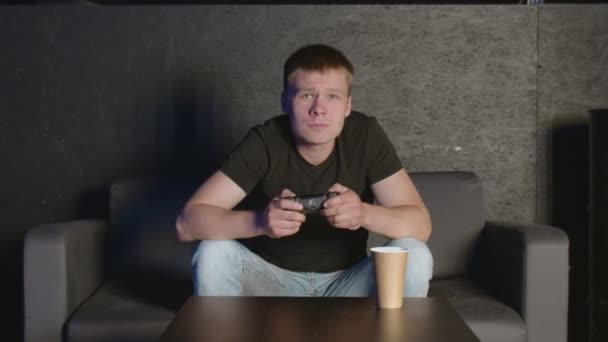 Młody człowiek gra w grę konsolową przed kamerą, pokonując przeciwnika online. Biały człowiek na kanapie. - Materiał filmowy, wideo