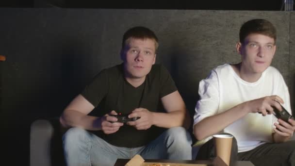 In de woonkamer Twee vrienden zitten op een bank videospel te spelen en pizza te eten. - Video