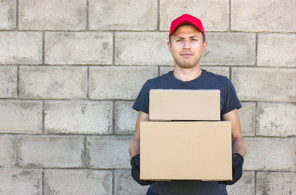 un jeune homme livreur se tient sur le fond d'un mur de pierre grise, tient deux boîtes en carton dans ses mains, dans un chapeau rouge et T-shirt gris, livraison, place pour une inscription - Photo, image
