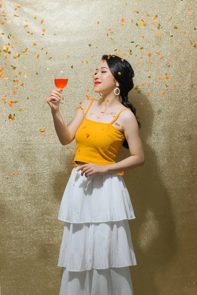Belle jeune femme avec confettis, célébration avec verre de champagne sur fond d'or. - Photo, image