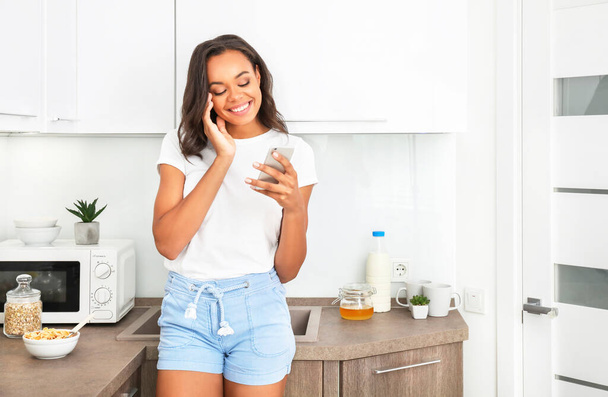 Young african woman eating corn flakes for breakfast in kitchen and talking on phone. - Photo, Image