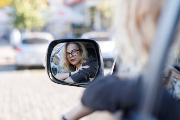 Belle jeune femme aux cheveux bouclés blonds portant un costume et des lunettes dans une voiture. Réflexion dans un miroir - Photo, image