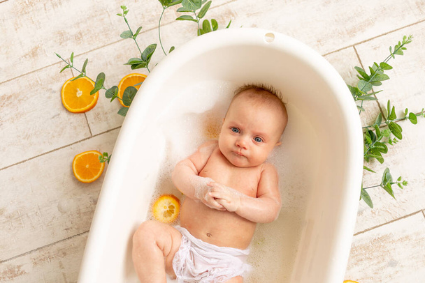 een klein kind baadt in een bad met schuim en sinaasappels, kinderen zijn allergisch voor citrus - Foto, afbeelding