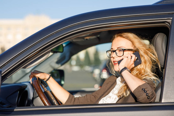 Belle jeune femme d'affaires aux cheveux bouclés blonds portant un costume et des lunettes dans une voiture parlant par téléphone portable - Photo, image