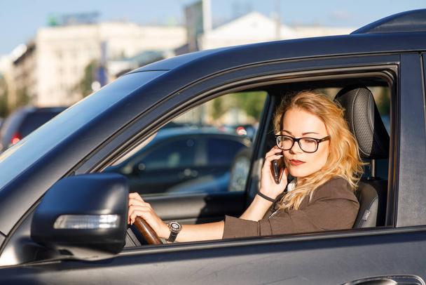 Όμορφη νεαρή επιχειρηματίας με ξανθά σγουρά μαλλιά φορώντας κοστούμι και γυαλιά σε ένα αυτοκίνητο μιλάμε από κινητό τηλέφωνο - Φωτογραφία, εικόνα