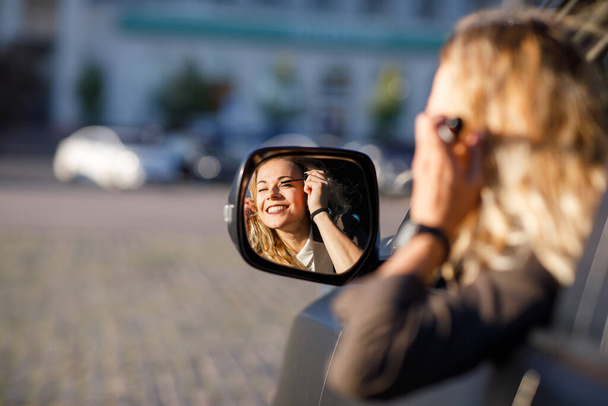Gyönyörű fiatal nő szőke göndör haj visel öltönyt és szemüveget pisztollyal egy autó szabadtéri mosolyog a tükörben egy autóban - Fotó, kép