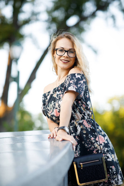 Beautiful young woman with blond curly hair wearing a dress and glasses  in a park in the evening - 写真・画像