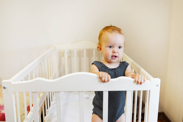 Beautiful baby standing in crib. Portrait of cute baby girl stand in cot and looking around. Toddler learning to stand in bed at home. - Photo, Image