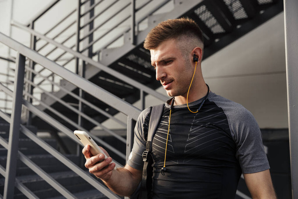 Image of young athletic sportsman with earphones using cellphone while working out near urban stair outdoors - 写真・画像