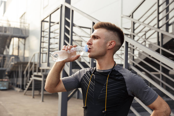 Image of unshaven caucasian sportsman drinking water while working out near urban stair outdoors - Photo, Image