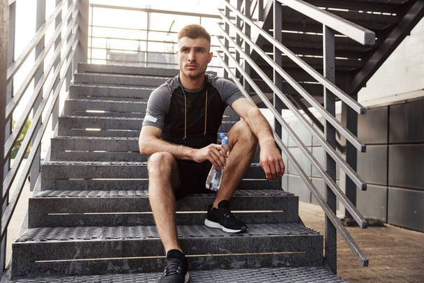 Image of unshaven caucasian sportsman drinking water while sitting on urban stair outdoors - Fotografie, Obrázek