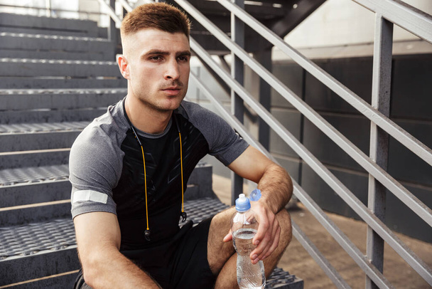 Image of unshaven caucasian sportsman drinking water while sitting on urban stair outdoors - Photo, Image