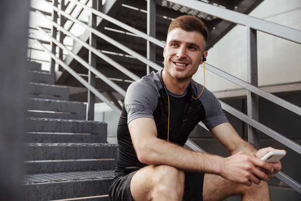 Image of laughing caucasian sportsman using cellphone and earphones while sitting on urban stair outdoors - Foto, immagini