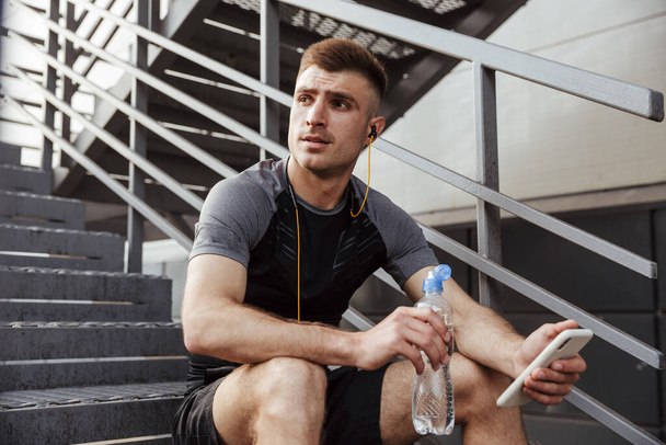 Image of unshaven caucasian sportsman drinking water and using cellphone while sitting on urban stair outdoors - Φωτογραφία, εικόνα