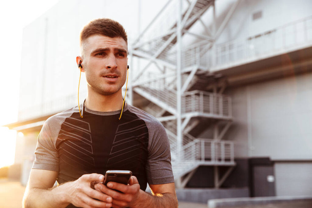 Image of young athletic sportsman using cellphone and earphones while working out at urban area outdoors - Foto, Imagen