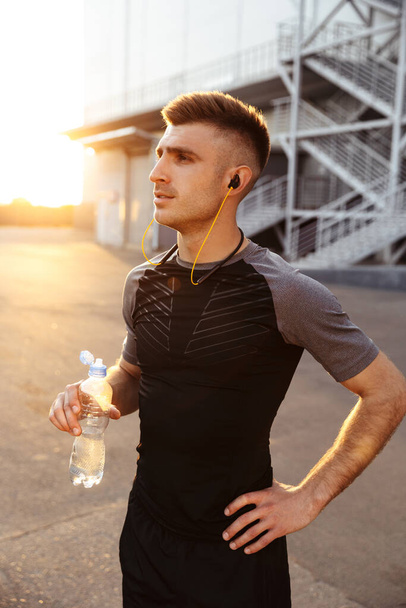 Image of young athletic sportsman in earphones drinking water while working out at urban area outdoors - Foto, Imagen