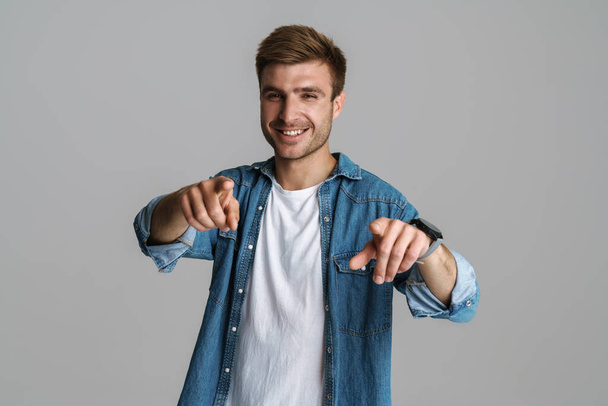Portrait of cheerful unshaven man laughing and pointing fingers at camera isolated over grey background - Foto, afbeelding
