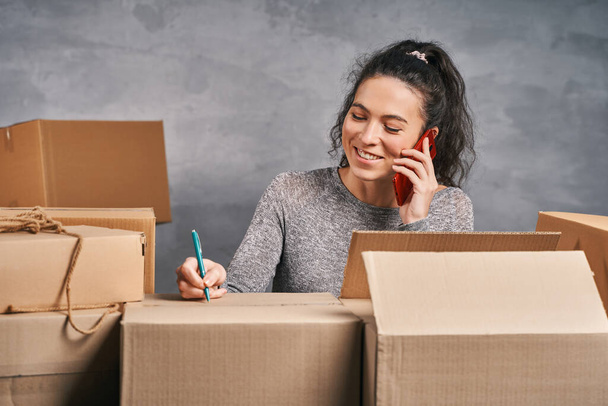 Жінка пише адреси на коробках, готує посилки для надсилання та отримання телефонних дзвінків. Робота з дому
 - Фото, зображення