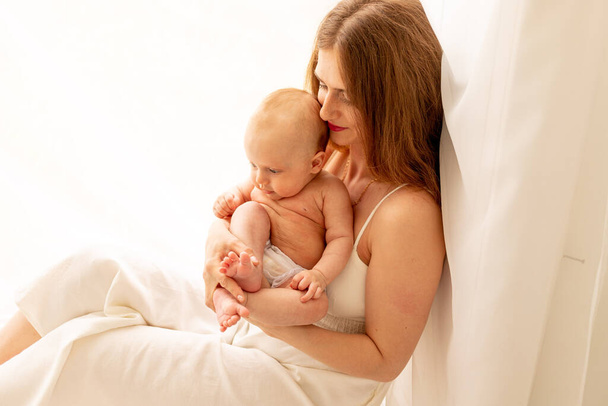 uma jovem mãe gentilmente abraça uma criança em seus braços enquanto está sentada na janela, o conceito de amor da mãe e cuidado com a criança. - Foto, Imagem