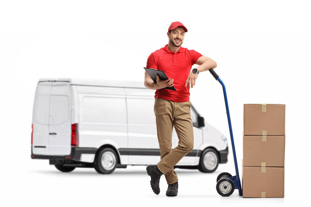 Full length portrait of a delivery man with cardboard boxes on hand truck standing in front of a white van isolated on white background - 写真・画像